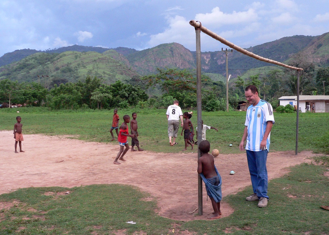 Barn som spelar fotboll på en grusplan på landsbygden med en vuxen i Argentinatröja, vilket illustrerar idrottens förmåga att förena människor.
